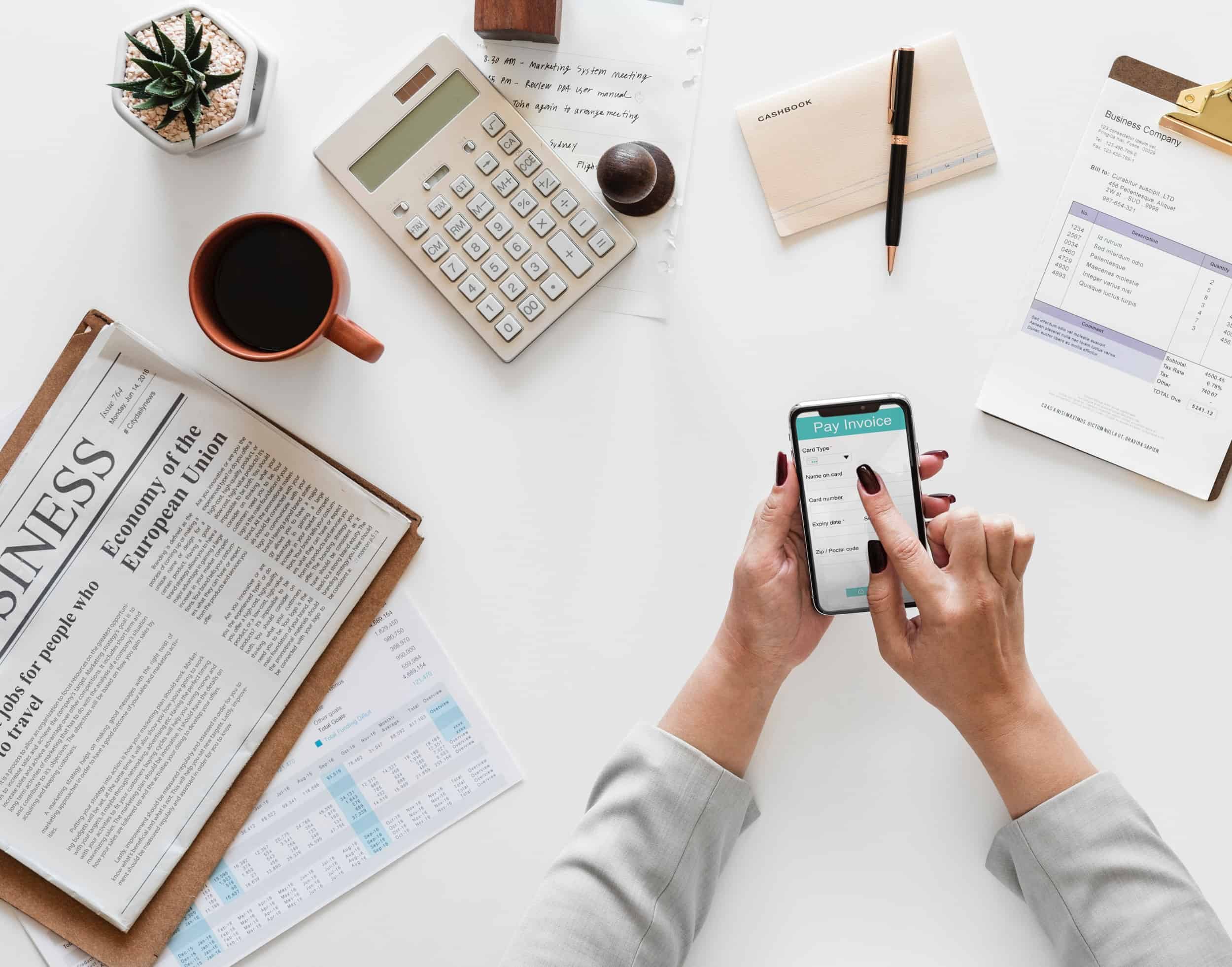 white desk with newspaper and coffee on it with someone making payment on her phone