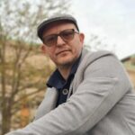 Portrait of Yannis Kotsinos, Software Engineer and AI Navigator at Zartis, photographed outdoors in a flat cap, tinted glasses and a grey wool coat.